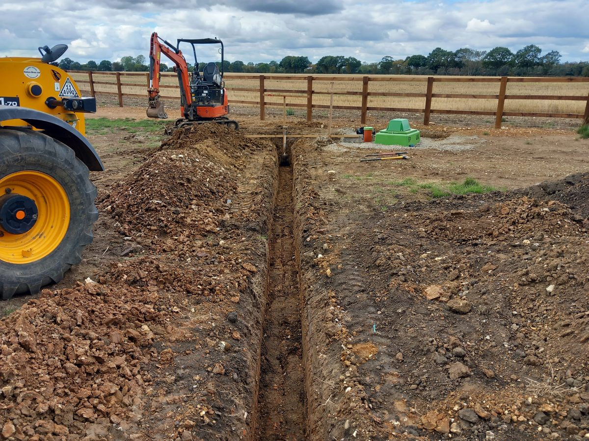 A narrow drainage trench dug across a field, with a mini digger and spoil alongside